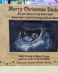 Christmas Baby Scan Photo Frame, reads Merry Christmas Daddy photo frame from bump, a brown wooden engraved photo frame on a sideboard against a blue wall standing beside a teddy bear with a baby scan photo insert. Baby in the scan is wearing a red and white santa hat by The Craft Collection Ireland