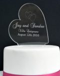 Heart Cake Topper shows a black background and a white iced cake with a pink ribbon wrapped around the bottom. On top of the cake stands a clear acrylic sideways heart engraved with two intertwined hearts and the words "Jay and Sandra Villa Catignano August 12th, 2010". Engraved personalised cake topper by The Craft Collection in Ireland