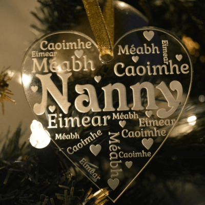 Grandparent Christmas Ornament features a clear acrylic glass-like heart ornament, see through with the word Nanny in the centre and first names surrounding it with little hearts dotted throughout engraved or etched appearing in white writing. Hanging from a gold glittery ribbon with a Christmas tree and lights in the background.