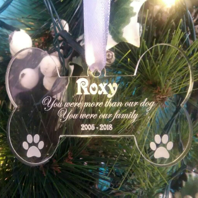 Dog Memorial Ornament, picture of a clear acrylic, glass like, dog bone christmas tree ornament,hanging on a silver ribbon against a lit christmas tree background that reads "Roxy, You were more the our dog, you were our family 2005-2018" by The Craft Collection Ireland